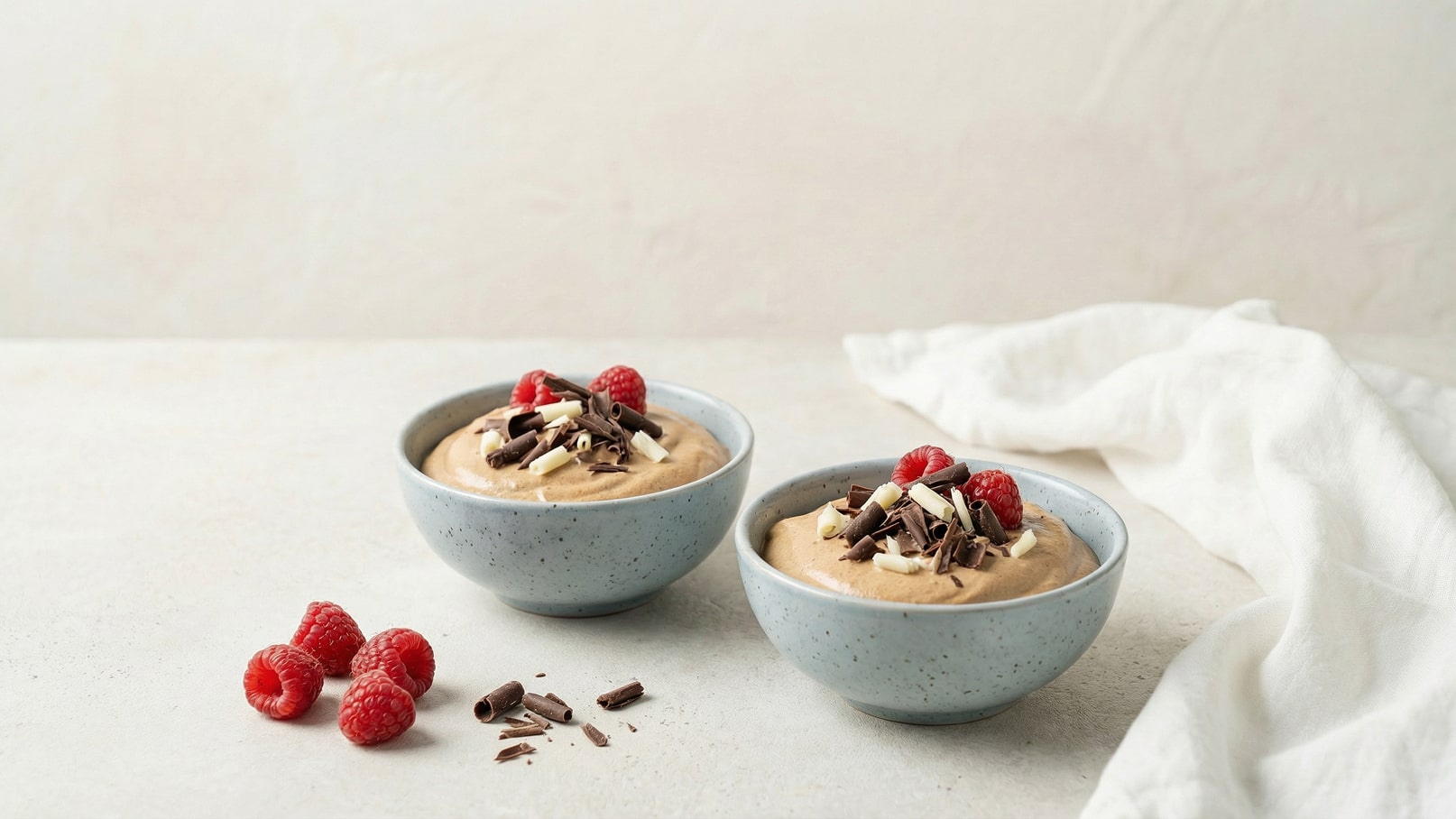 Zwei Schalen mit cremigem Dessert sind auf einem hellen Tisch platziert. Jede Schale ist mit Himbeeren und Schokoladenspänen dekoriert. Neben den Schalen liegen einige frische Himbeeren und Schokoladenstückchen.