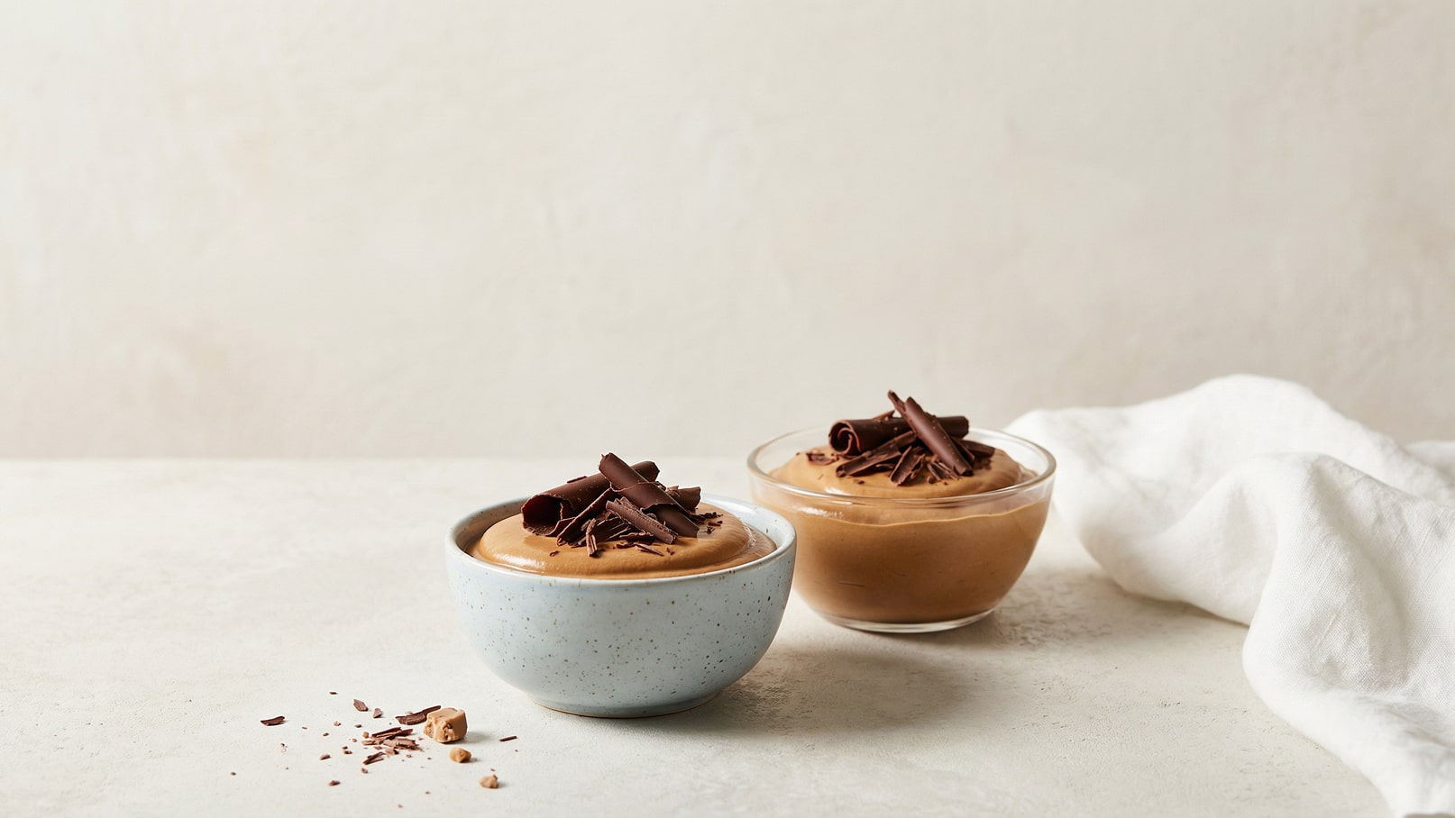 Zwei Schalen mit Schokoladenmousse, eine in hellblau und eine transparent, beide mit Schokoladenraspeln dekoriert. Im Hintergrund ist ein leichter, schlichter Untergrund sichtbar.