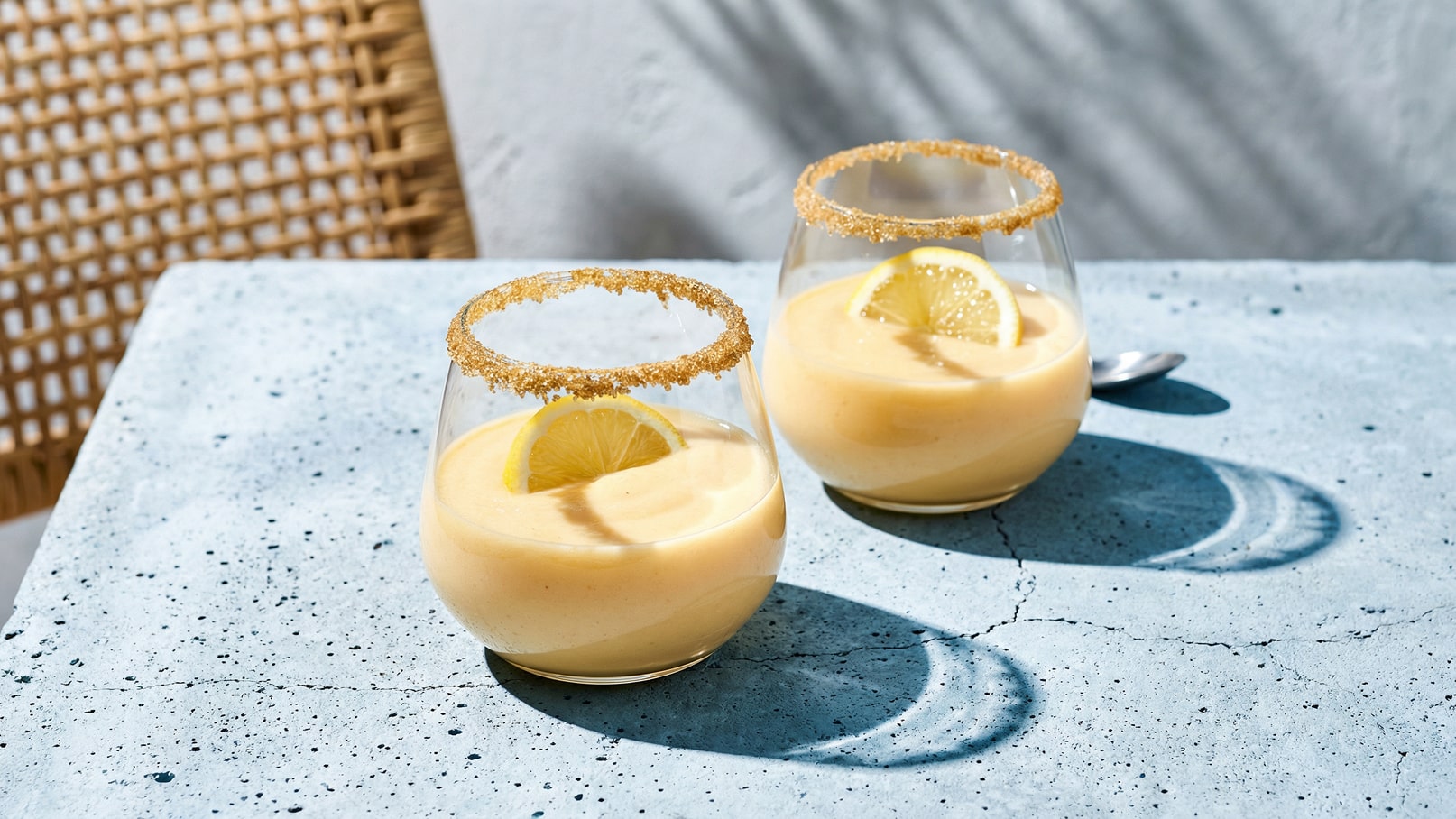 Zwei Gläser mit einem cremigen, gelben Dessert stehen auf einem hellen Tisch. Jedes Glas hat einen Rand aus braunem Zucker und ist mit einer Zitronenscheibe garniert. Die Szenerie vermittelt eine frische, einladende Atmosphäre.