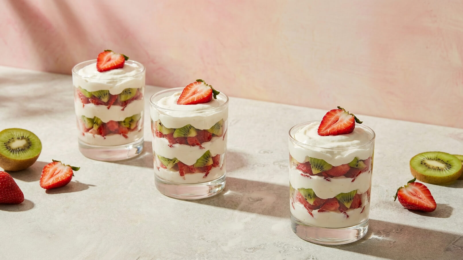 Drei durchsichtige Gläser mit Schichten aus Joghurt, Erdbeeren und Kiwi. Jedes Glas ist mit einer Schicht Sahne bedeckt und mit einer Erdbeere garniert. Einige Erdbeeren und eine Kiwi liegen auf dem Tisch neben den Gläsern. Der Hintergrund ist in sanften rosa Tönen gehalten.