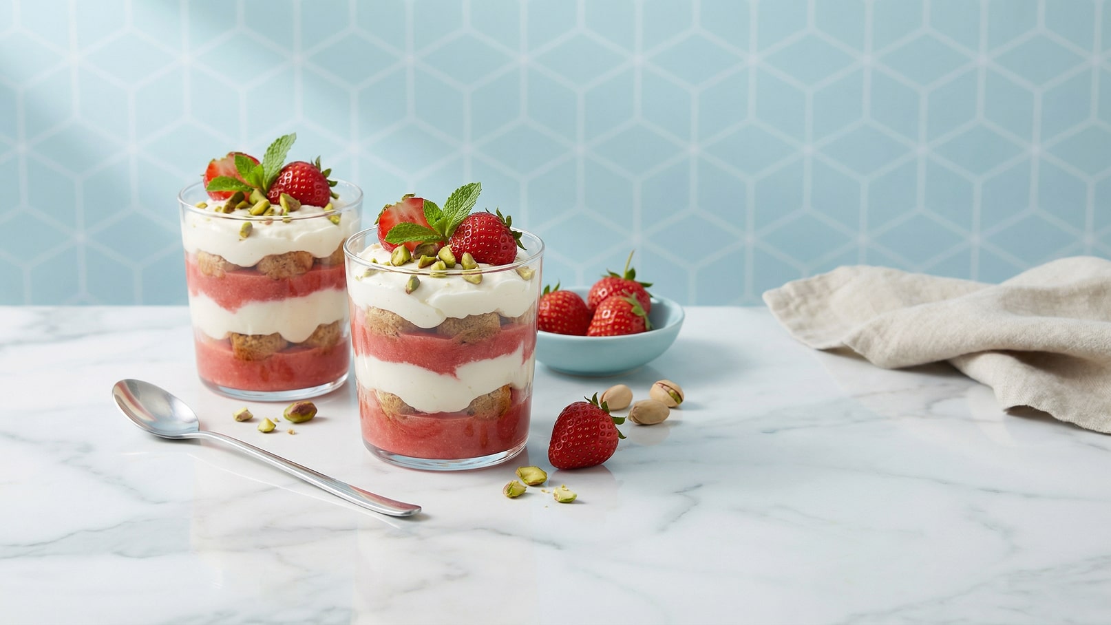 Zwei Gläser mit Schichtdessert, gefüllt mit Joghurt, Erdbeeren und knusprigen Keksen, garniert mit gehackten Pistazien und frischen Erdbeeren. Im Hintergrund ein kleiner Teller mit weiteren Erdbeeren. Die Oberfläche ist marmoriert und hell.