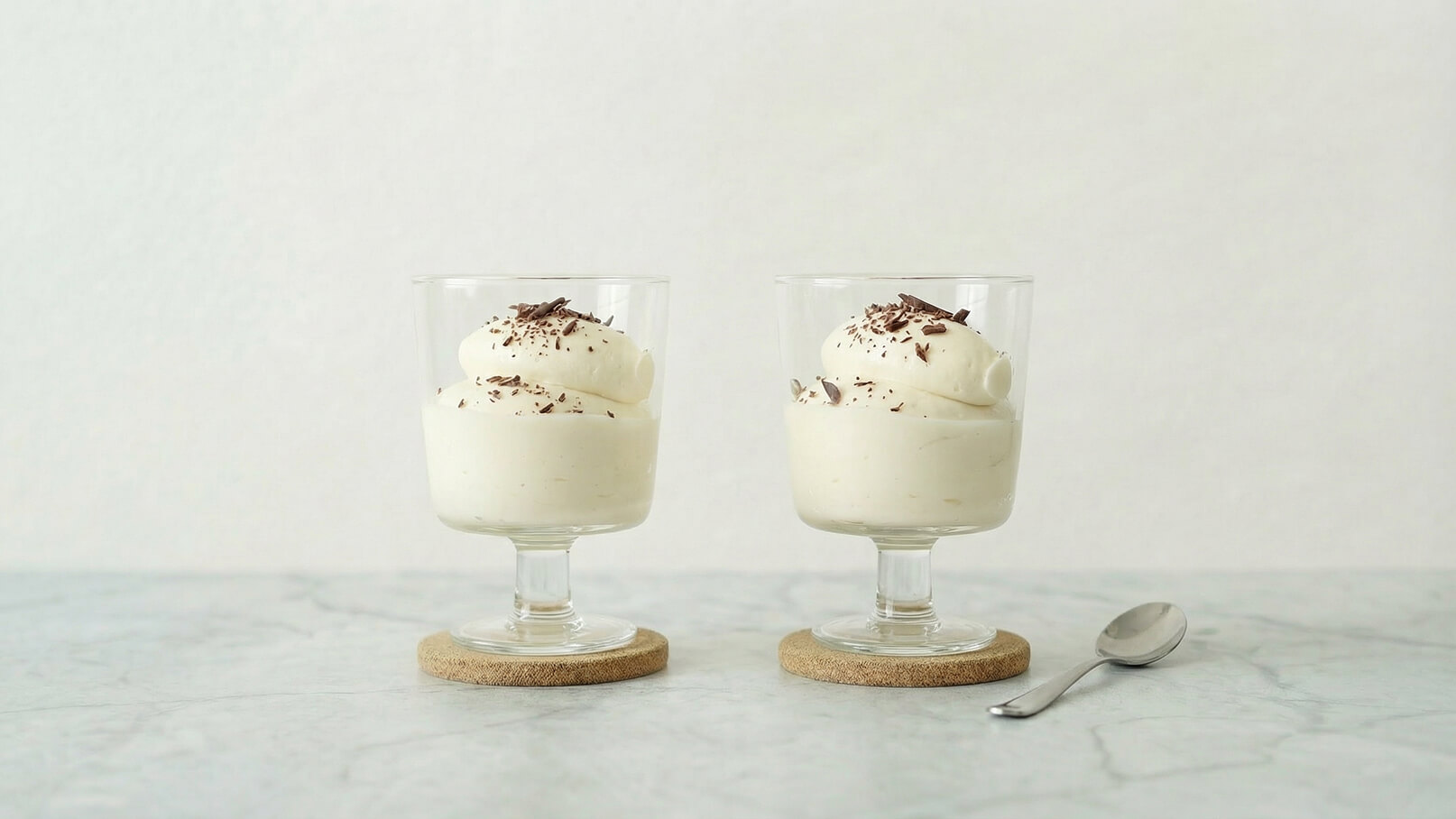 Zwei Gläser mit weißem Sahne-Dessert stehen auf einem Tisch. Jedes Glas ist mit einer Haube aus Schlagrahm und Schokoladenspänen dekoriert. Daneben liegt ein kleiner Löffel. Der Hintergrund ist schlicht und hell.