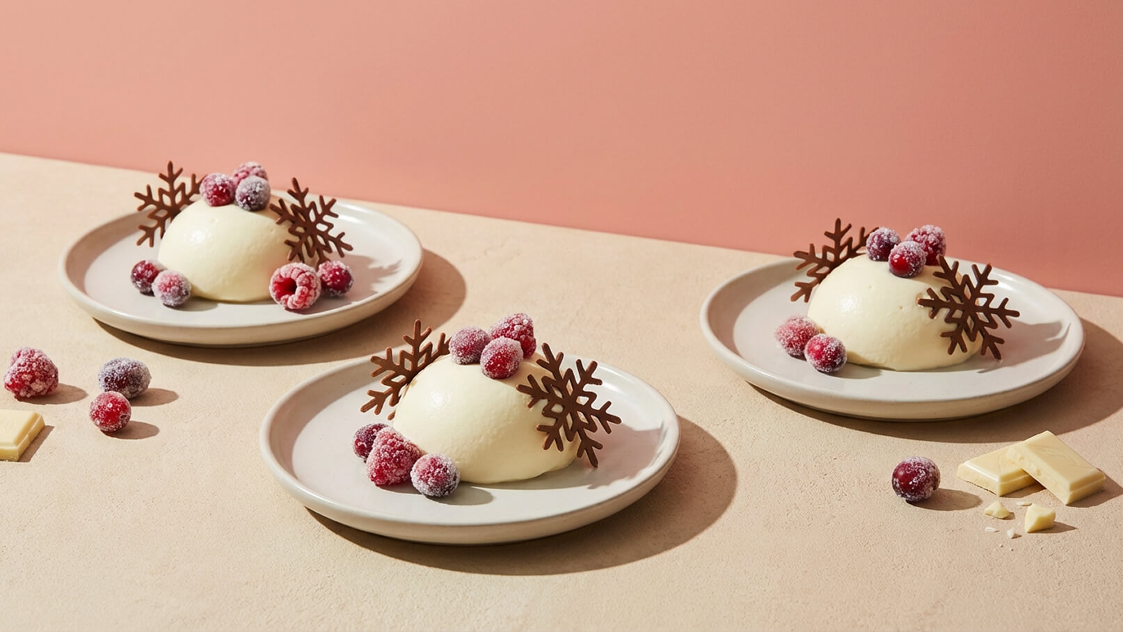 Drei Teller mit einer cremigen Dessertkugel, dekoriert mit gefrorenen Himbeeren und schokoladigen Schneeflocken. Eine Portion ist leicht nach vorne geneigt, während die anderen zwei daneben stehen. Im Hintergrund ist eine sanfte rosa Wand sichtbar.