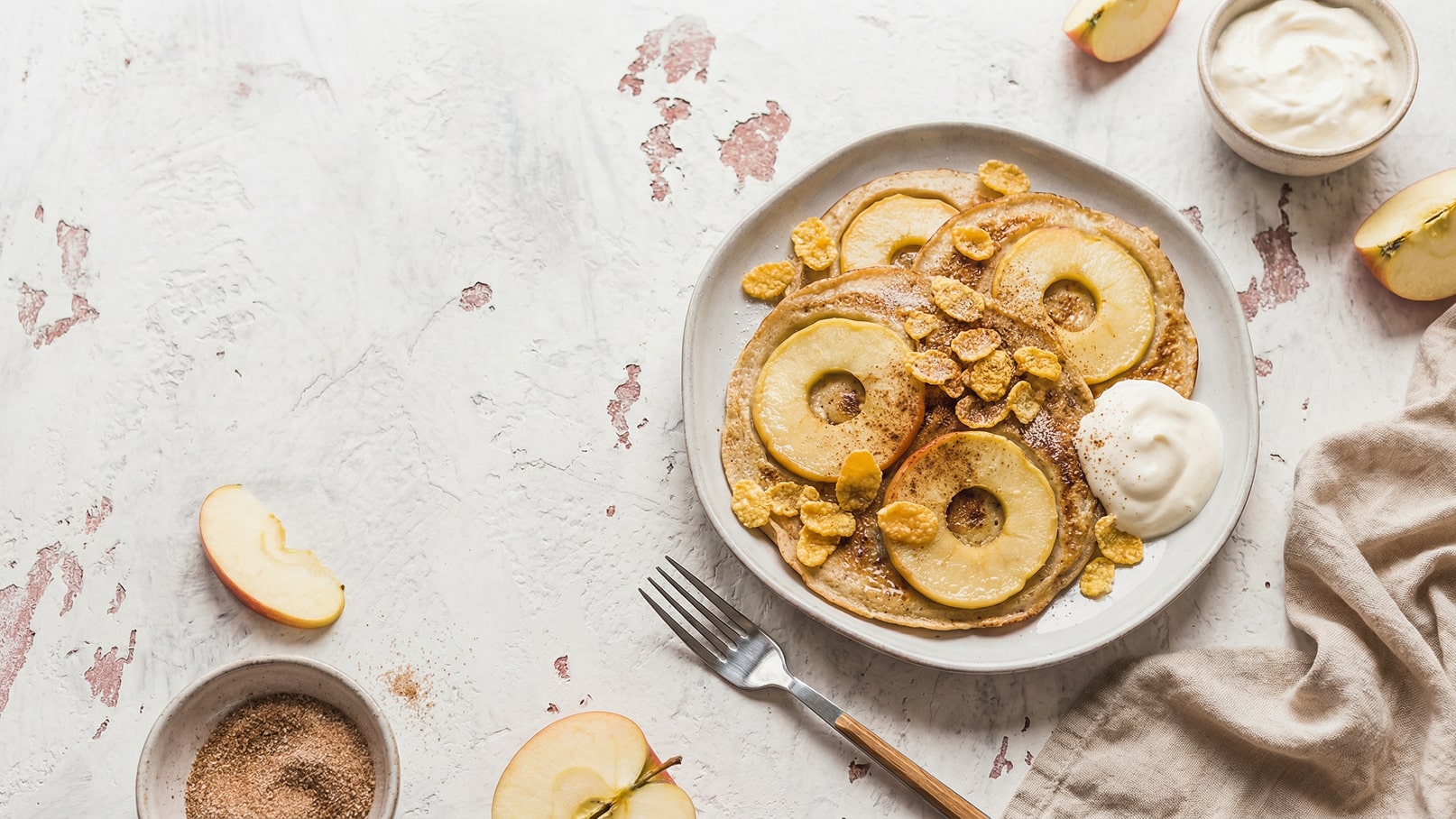 Ein Teller mit Apfelpfannkuchen, auf denen Apfelringe und Cornflakes liegen. Daneben befindet sich ein Schälchen mit Joghurt und ein halber Apfel. Die Präsentation vermittelt einen frischen und einladenden Frühstückslook.