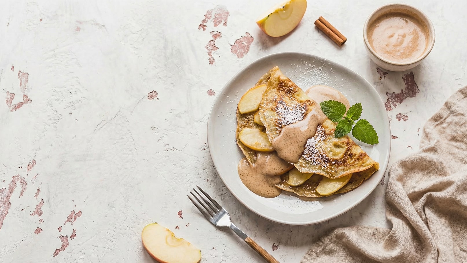 Ein Teller mit gefüllten Pfannkuchen, die mit Apfelscheiben bedeckt sind, steht auf einem hellen Tisch. Darauf ist eine cremige Soße und Puderzucker zu sehen. Frische Minze und eine Gabel ergänzen das Bild. Ein Apfelstück und Zimtstangen liegen neben dem Teller.