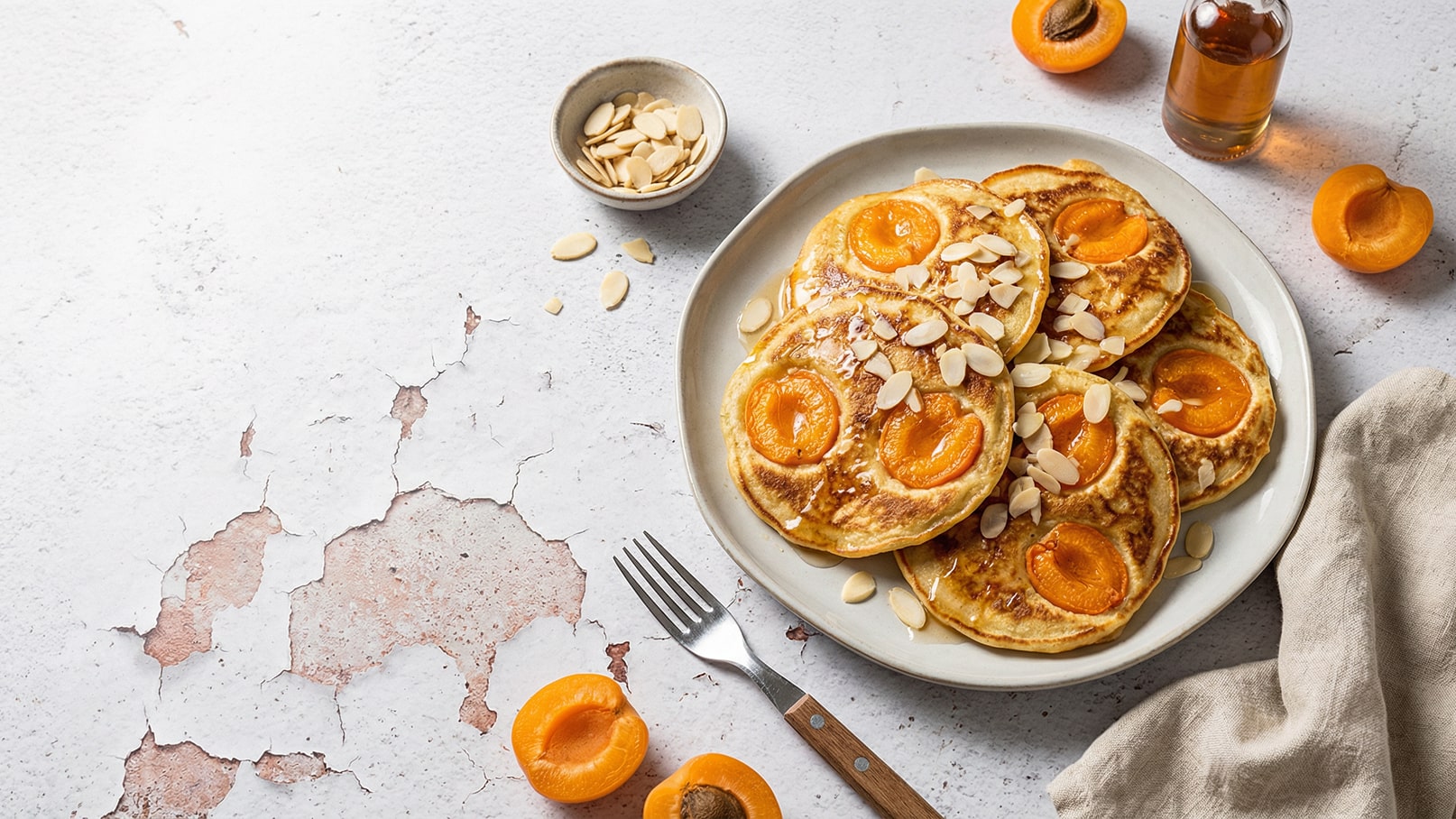 Ein Teller mit goldbraunen Pfannkuchen, belegt mit halbierten Aprikosen und gehobelten Mandeln. Daneben befinden sich eine kleine Schüssel mit Mandeln und einige frische Aprikosen. Ein Fläschchen mit Sirup steht ebenfalls auf dem Tisch.