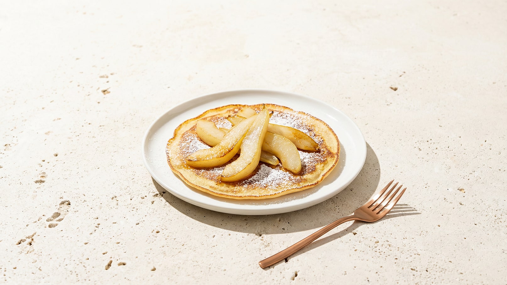 Ein flacher Pfannkuchen liegt auf einem weißen Teller, garniert mit karamellisierten Birnenscheiben und einem Hauch Puderzucker. Daneben liegt eine rosa-goldene Gabel. Der Hintergrund ist ein heller, strukturierter Tisch.