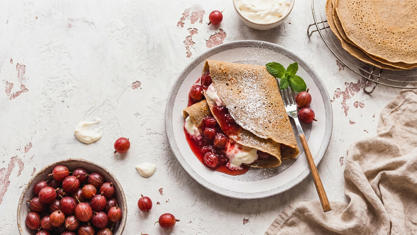 Ein Teller mit einer gefüllten Crepe, die mit roten Johannisbeeren und Sahne serviert wird. Eine Gabel mit einem Holzgriff liegt daneben. Im Hintergrund befinden sich weitere Crepes und eine Schale mit roten Johannisbeeren.