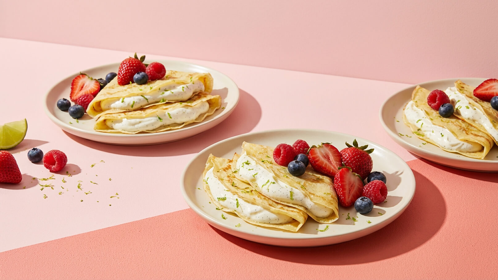 Drei Teller mit gefüllten Crêpes sind vor einem rosa Hintergrund angeordnet. Die Crêpes sind mit Sahne gefüllt und mit Limettenschale bestreut. Frische Erdbeeren, Himbeeren und Heidelbeeren liegen dekorativ auf den Tellern.