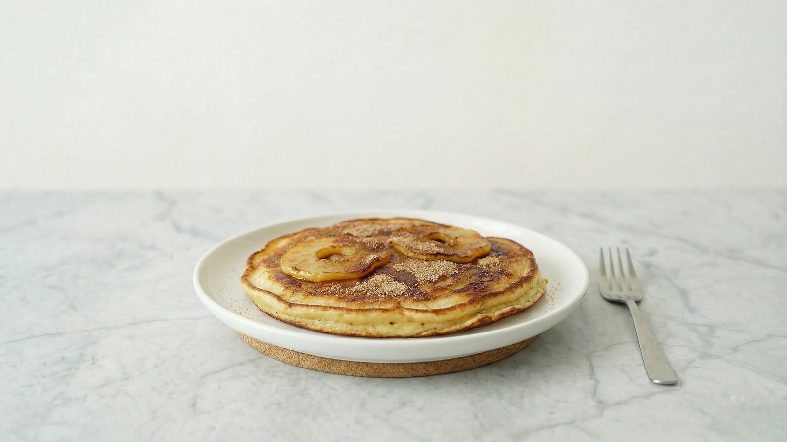 Ein weißer Teller mit einem Stapel von Pfannkuchen, auf denen gebratene Bananenscheiben liegen. Die Pfannkuchen sind leicht goldbraun und mit einer Prise Zimt bestäubt. Der Teller steht auf einem Holzteller, neben dem eine Gabel liegt.