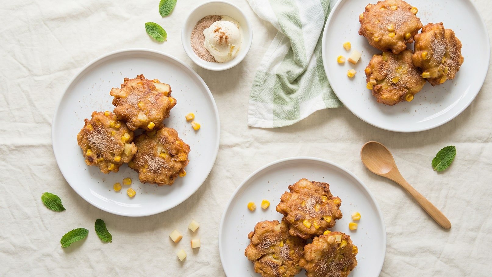 Auf den Tellern liegen mehrere goldbraune Maispfannkuchen, bestreut mit Zucker und Zimt. Neben den Pfannkuchen ist eine kleine Schale mit einer Kugel Eis. Die Anrichte erfolgt auf einem hellen Tischtuch, gesäumt von einigen frischen Minzblättchen.