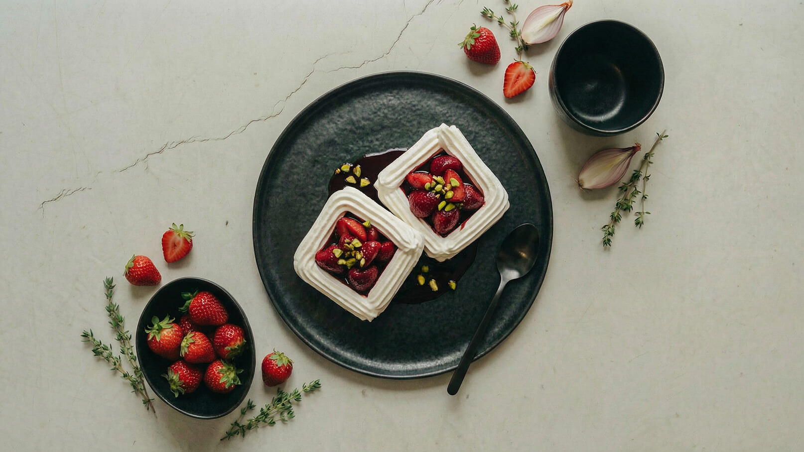 Zwei quadratische Desserts mit Sahne, frischen Erdbeeren und gehackten Pistazien auf einem schwarzen Teller, umgeben von Erdbeeren und Kräutern. Ein Löffel liegt neben dem Teller.
