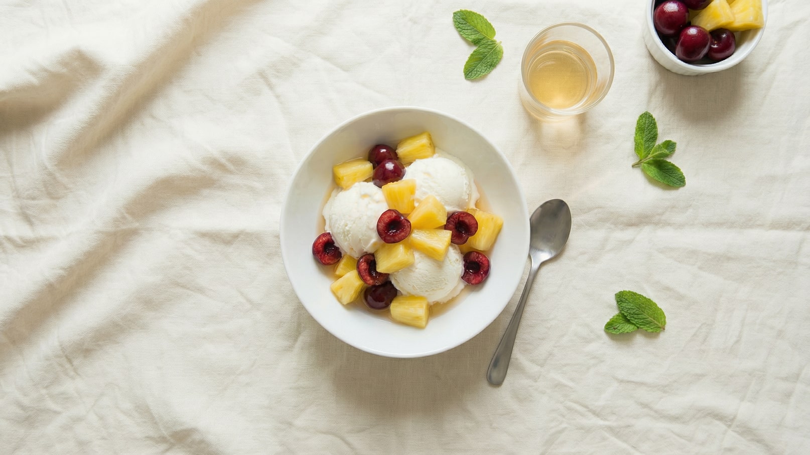 Eine weiße Schüssel mit drei Kugeln Vanilleeis, dekoriert mit Ananasscheiben und roten Kirschen. Daneben ein kleiner Löffel und ein Glas mit einem hellen Getränk. Frische Minzblätter liegen auf der Tischdecke.