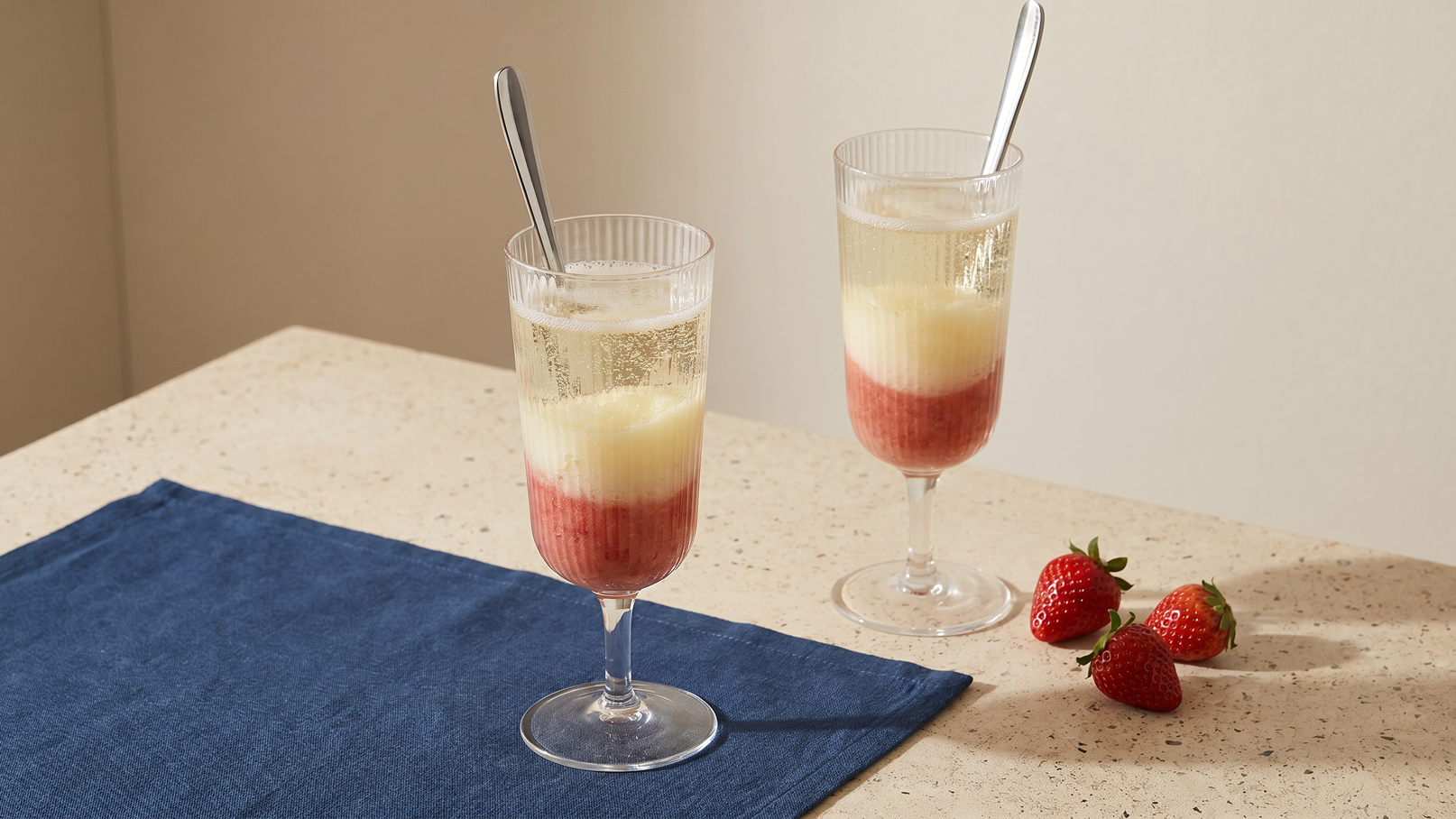 Zwei hohe Gläser mit Schaumwein und einer Schicht aus Erdbeerpüree stehen auf einem Tisch. Neben den Gläsern liegen frische Erdbeeren und ein blauer Stoffuntersetzer ist sichtbar.