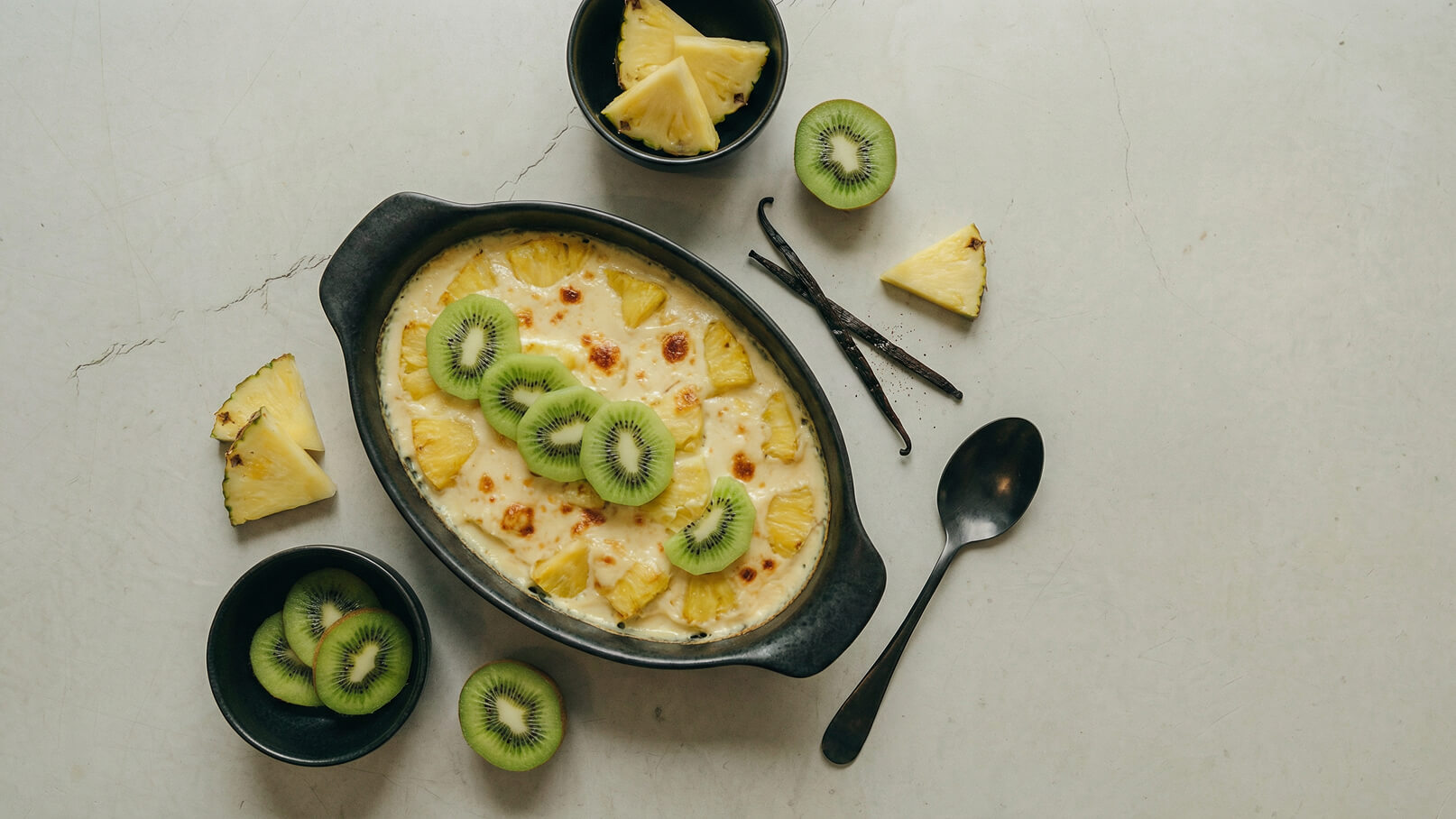Ein schwarzes Backgeschirr mit einer cremigen Mischung aus Ananas und Kiwi. Darüber liegen Scheiben von Kiwi und Ananas. Daneben sind frische Kiwis und Ananasstücke sowie zwei Vanilleschoten. Ein Löffel liegt auf dem Tisch.