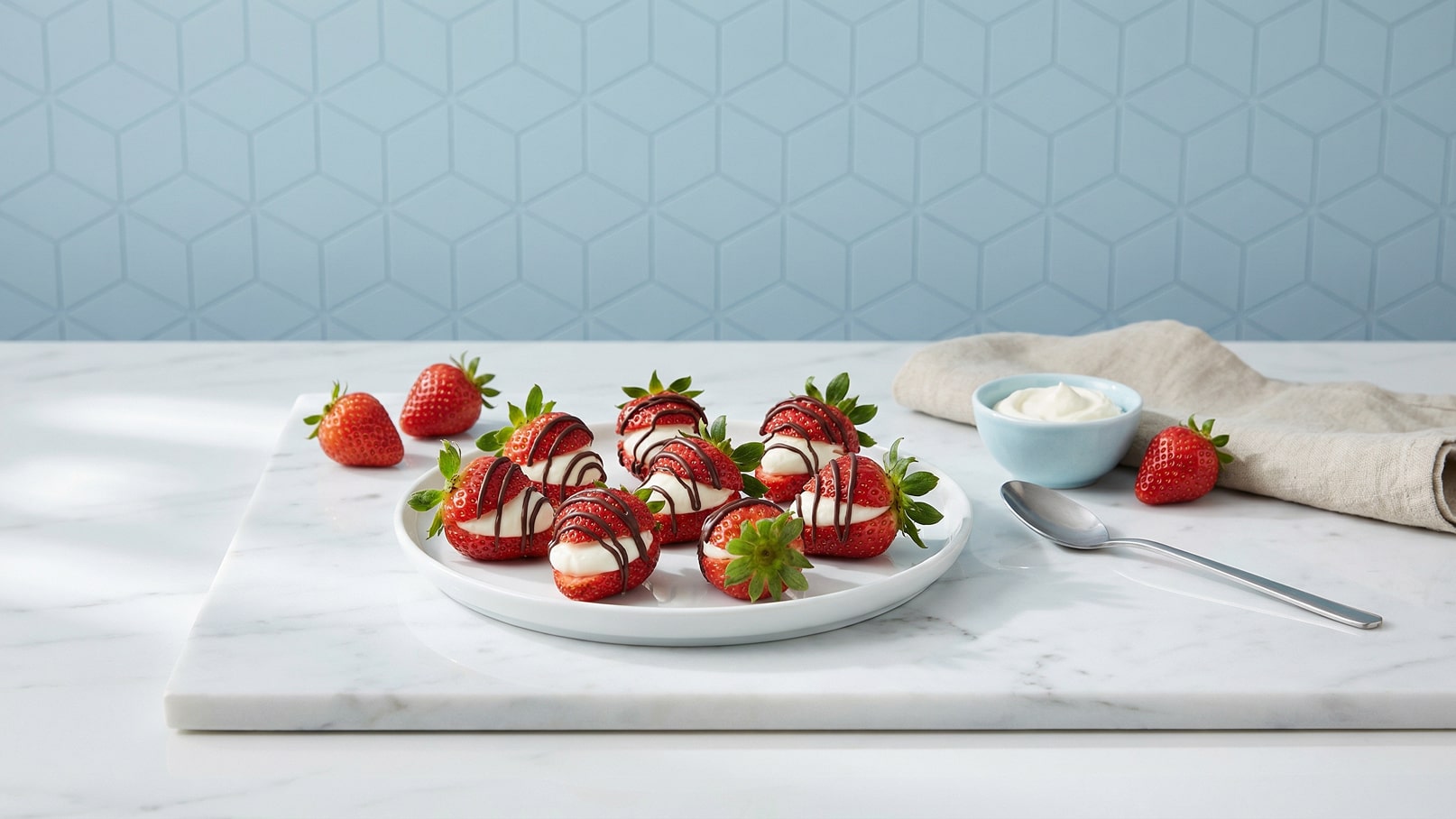 Ein weißer Teller mit gefüllten Erdbeeren, die mit Schlagsahne und Schokolade verziert sind, steht auf einem marmorartigen Untergrund. Neben dem Teller liegen frische Erdbeeren, ein silberner Löffel und eine kleine Schüssel mit weiteren Schlagsahne. Der Hintergrund ist in sanftem Blau.