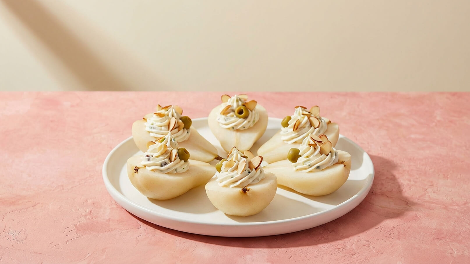 Sechs gefüllte Birnenhälften auf einem weißen Teller. Jede Birne ist mit cremiger Füllung dekoriert, die mit Mandelscheiben und einer Olive garniert ist. Der Teller steht auf einer rosa Oberfläche.