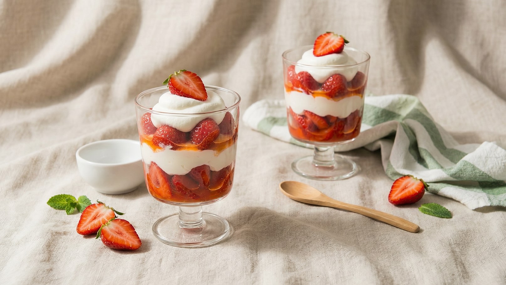 Zwei Gläser mit Erdbeerdessert stehen auf einer hellen Stoffunterlage. Jedes Glas enthält Schichten aus Erdbeeren, Sahne und einer fruchtigen Soße, gekrönt mit frischen Erdbeeren. Daneben liegt ein kleiner Holzlöffel und daneben einige halbierte Erdbeeren sowie ein frisches Minzblatt.