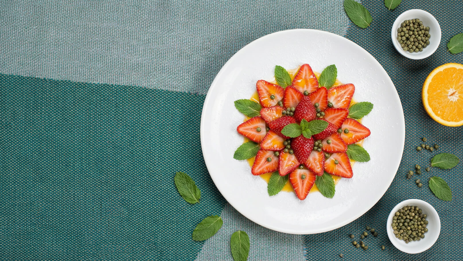 Ein rundes weißes Teller mit kunstvoll angerichteten Erdbeeren in Form einer Blüte, umgeben von frischen Minzblättern. Im Hintergrund liegen kleine Schalen mit grünen Pfefferkörnern und eine halbe Orange. Der Tisch ist mit einem strukturierten, grün-blauen Tuch bedeckt.