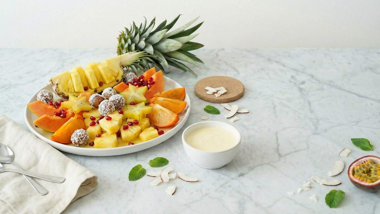 Auf dem Bild ist ein frischer Obstteller mit Ananas, Papaya, Sternfrucht und Granatapfelkernen zu sehen. Neben dem Teller steht eine kleine Schüssel mit einer cremigen Sauce und etwas Kokosnuss. Die Ananas liegt halbiert auf dem Teller, während ein Blatt Minze daneben liegt.