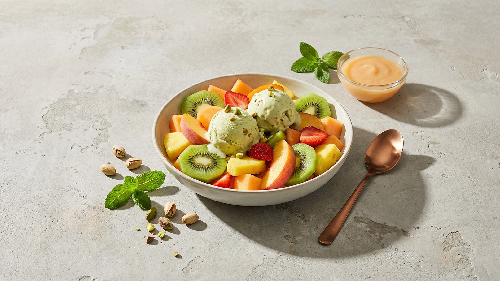 Eine Schüssel mit frischem Obst, darunter Melone, Kiwi, Erdbeeren und Ananas, ist mit zwei Kugeln Pistazieneis garniert. Neben der Schüssel befindet sich ein kleiner Behälter mit einer hellen Soße und ein Löffel aus Kupfer. Frische Minzblätter und einige Pistazien sind ebenfalls im Bild.