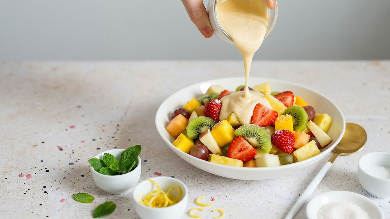 Eine Hand gießt eine cremige Sauce über eine bunte Schüssel mit frischem Obst, darunter Kiwi, Erdbeeren, Ananas und Trauben. Die Fruchtmischung ist appetitlich angerichtet und wirkt frisch und einladend.