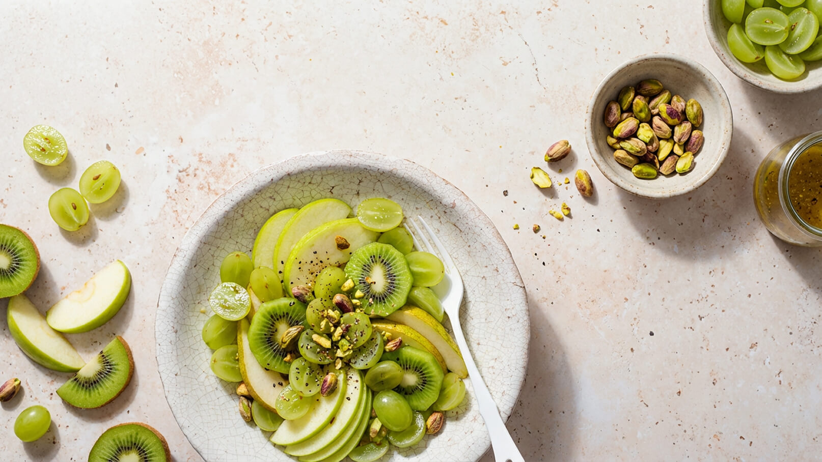 Eine Schüssel mit einem frischen Obstsalat aus grünen Äpfeln, Kiwis und grünen Trauben, garniert mit gehackten Pistazien. Eine weiße Gabel liegt neben der Schüssel. Im Hintergrund sind weitere Trauben und eine kleine Schale mit Pistazien zu sehen.