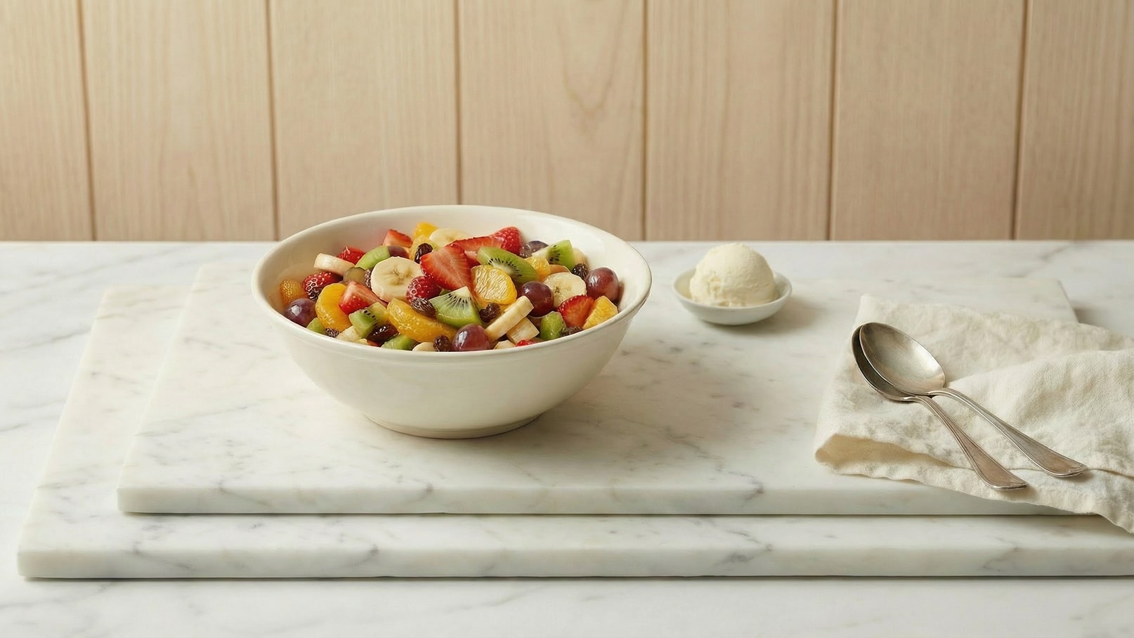Eine Schüssel mit einer bunten Obstsalat-Mischung, darunter Trauben, Bananen, Erdbeeren, Kiwi und Orangen, steht auf einer Marmoroberfläche. Neben der Schüssel befindet sich eine kleine Portion Vanilleeis und zwei Löffel liegen auf einer Serviette.