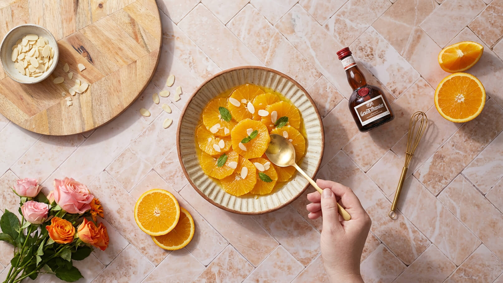 Eine Hand hält einen goldenen Löffel und schöpft aus einer Schüssel mit orangefarbenen Orangenscheiben, dekoriert mit Mandelsplittern und Minzblättern. Neben der Schüssel liegen frische Orangen, ein kleiner Behälter mit Mandeln und eine Flasche Likör.