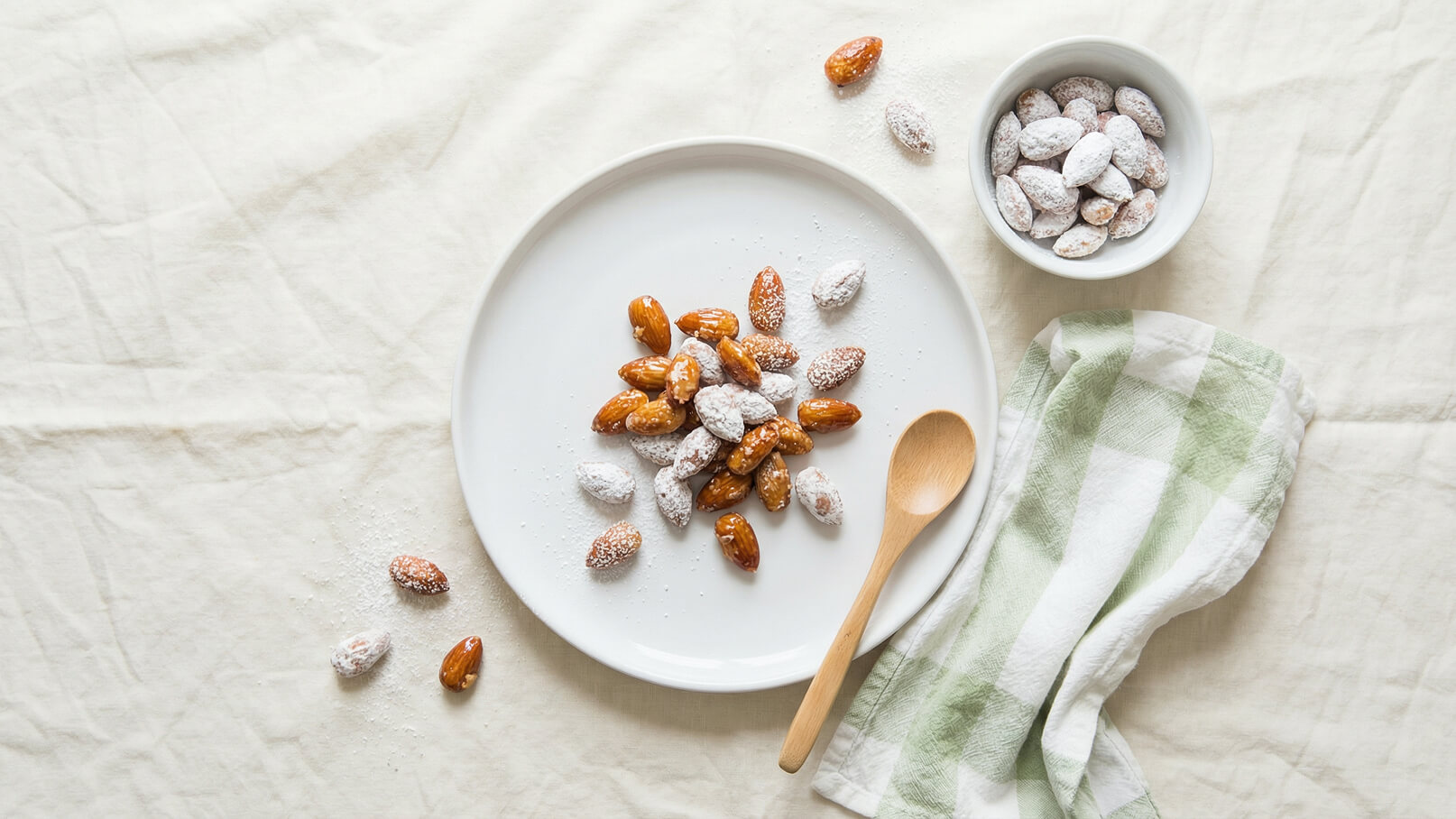 Ein weißer Teller mit einer Auswahl an süßen, mit Puderzucker bestäubten Mandeln und einer kleinen Schale mit ähnlichen Mandeln. Eine Holzlöffel liegt daneben, und ein grün-weiß kariertes Geschirrtuch ist ebenfalls sichtbar. Der Hintergrund ist schlicht und hell.