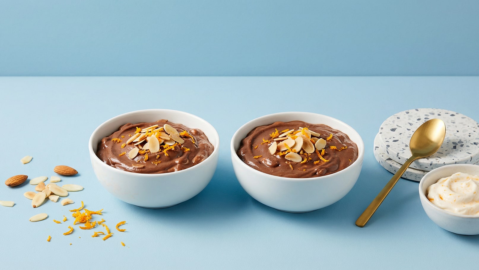 Zwei weiße Schalen mit cremigem Schokoladen-Dessert, garniert mit Mandelblättchen und orangener Schale. Daneben liegen einige ganze Mandeln, eine kleine Schüssel mit einer hellen Creme und ein goldener Löffel. Der Hintergrund ist in einem sanften Blau gehalten.
