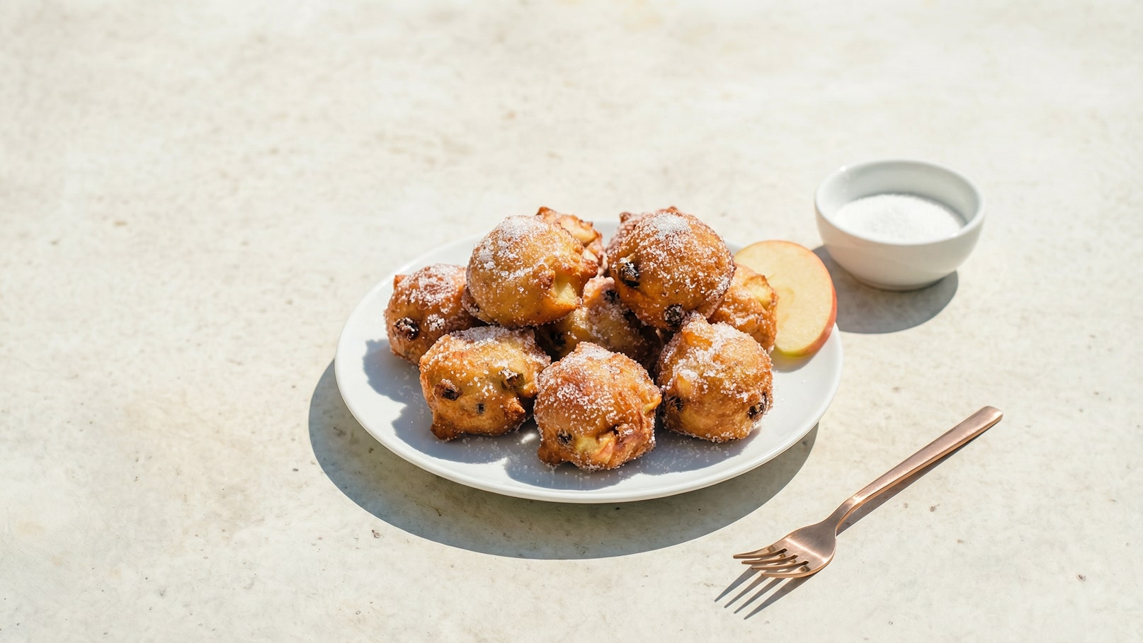 Ein Teller mit frisch gebackenen, goldbraunen Apfelküchle, bestäubt mit Puderzucker. Neben dem Teller liegt ein Stück Apfel und eine kleine Schüssel mit Zucker. Ein goldfarbener Gabel liegt ebenfalls daneben.