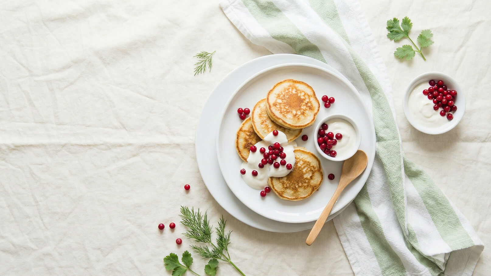Ein Teller mit kleinen Pfannkuchen, garniert mit roten Beeren und Sahne. Neben dem Teller steht eine kleine Schüssel mit weiteren roten Beeren und Sahne. Im Hintergrund liegt ein gestreiftes Tuch.