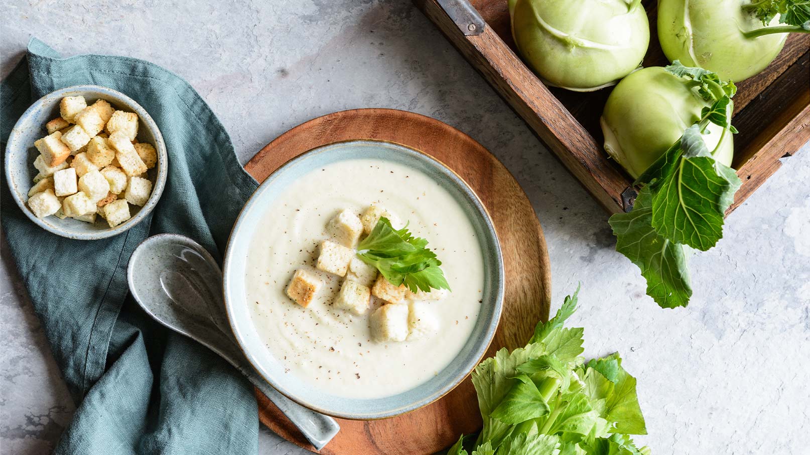 Auf dem Bild befindet sich eine Schüssel, gefüllt mit einer köstlichen Kohlrabisuppe. Als Topping sind Brotbrösel und frische Kräuter darauf drapiert, was dem Gericht eine ansprechende Optik verleiht. Die Schüssel steht auf einem rustikalen Holzbrett, das eine natürliche und gemütliche Atmosphäre schafft. Neben der Schüssel befindet sich eine weitere Schüssel mit Brotbröseln und ein türkises Tuch. Auf der anderen Seite steht eine Kiste mit frischem Kohlrabi, die die frischen Zutaten des Gerichts betont. 