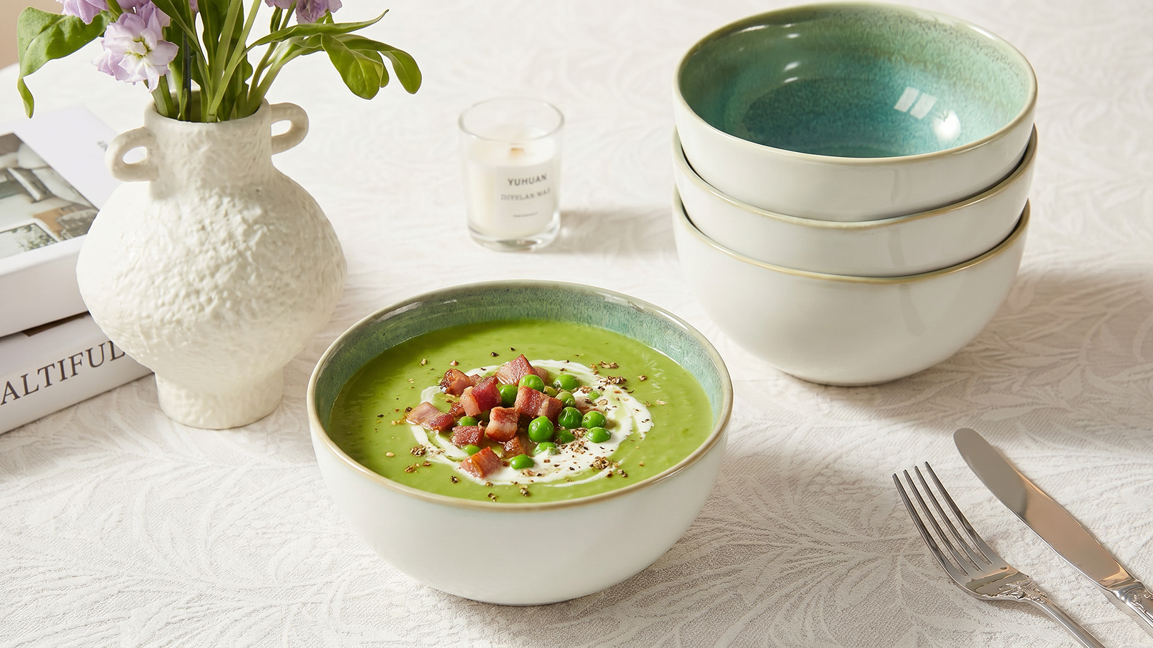 Eine Schüssel mit grüner Suppe, garniert mit Erbsen und gewürfeltem Schinken, steht auf einem Tisch. Daneben sind drei weitere Schalen gestapelt. Im Hintergrund befindet sich eine Vase mit Blumen und eine Kerze.