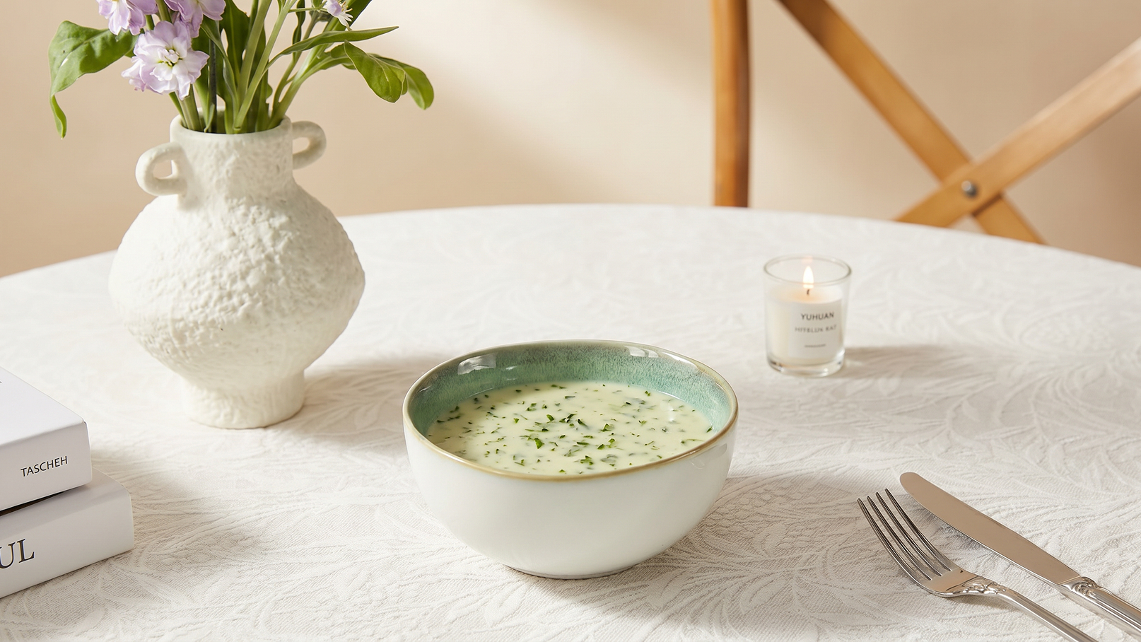 Eine weiße Schüssel mit cremiger Suppe, garniert mit frischen Kräutern, steht auf einem Tisch. Daneben befindet sich eine Vase mit bunten Blumen und eine kleine Kerze. Der Tisch ist mit einer eleganten Tischdecke bedeckt und es liegen Besteck und Bücher in der Nähe.