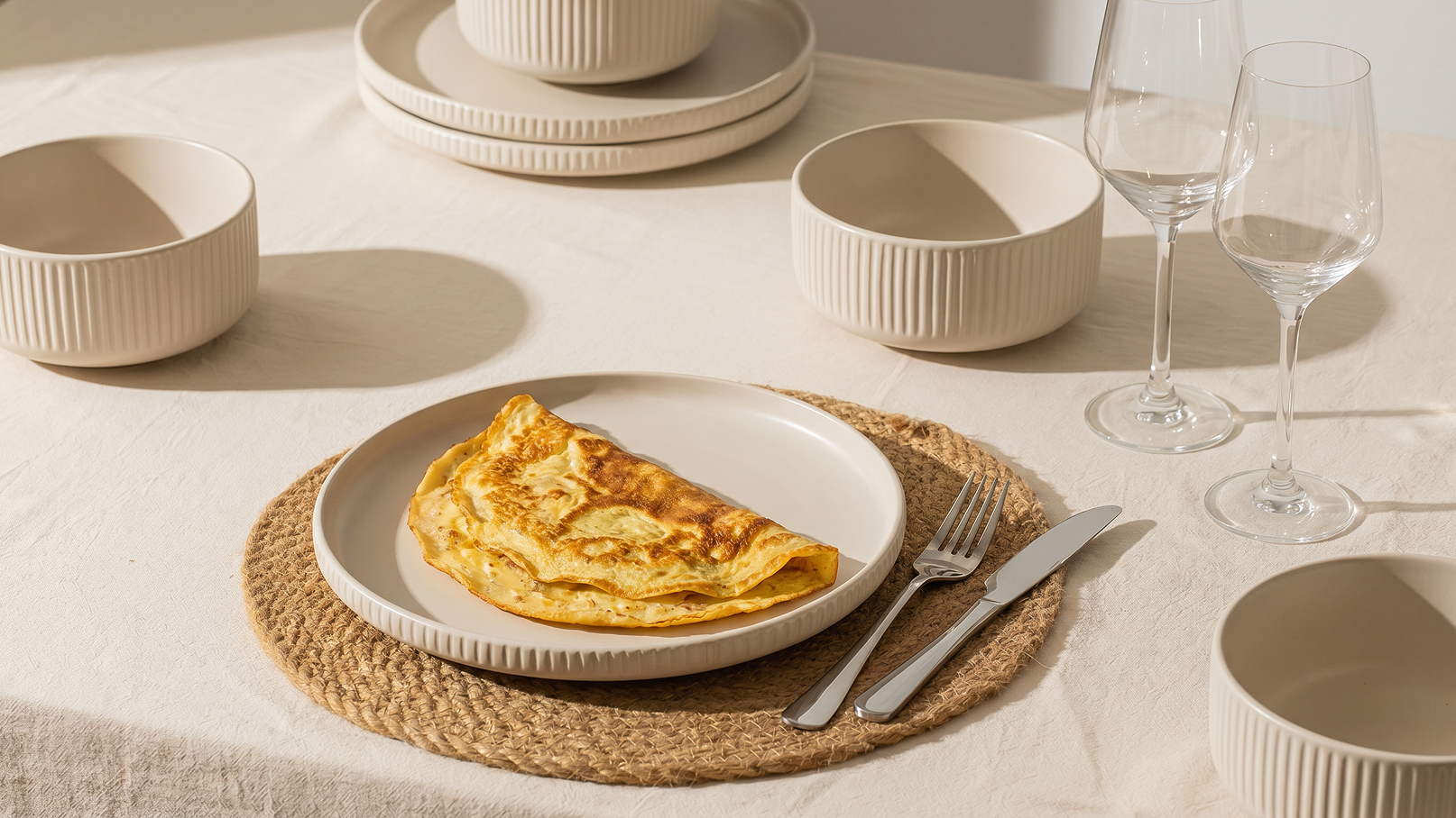 Auf dem Tisch liegt ein Teller mit einer goldbraunen Omelette, umgeben von neutralfarbenem Geschirr und Gläsern. Der Tisch ist mit einer hellen Tischdecke und einem runden Untersetzer dekoriert. Die Atmosphäre wirkt einladend und gemütlich.