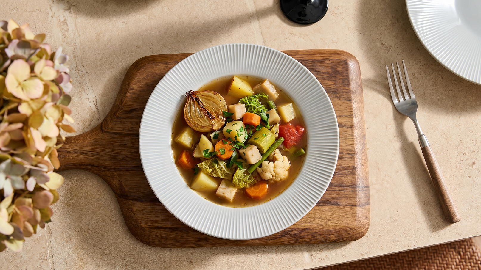 Eine Schüssel mit bunter Gemüsesuppe liegt auf einem Holzbrett. Die Suppe enthält Kartoffeln, Karotten, Blumenkohl und verschiedene grüne Gemüsesorten. Sie wird von einem weißen Rand umgeben und ist in der Mitte der Bildkomposition platziert.