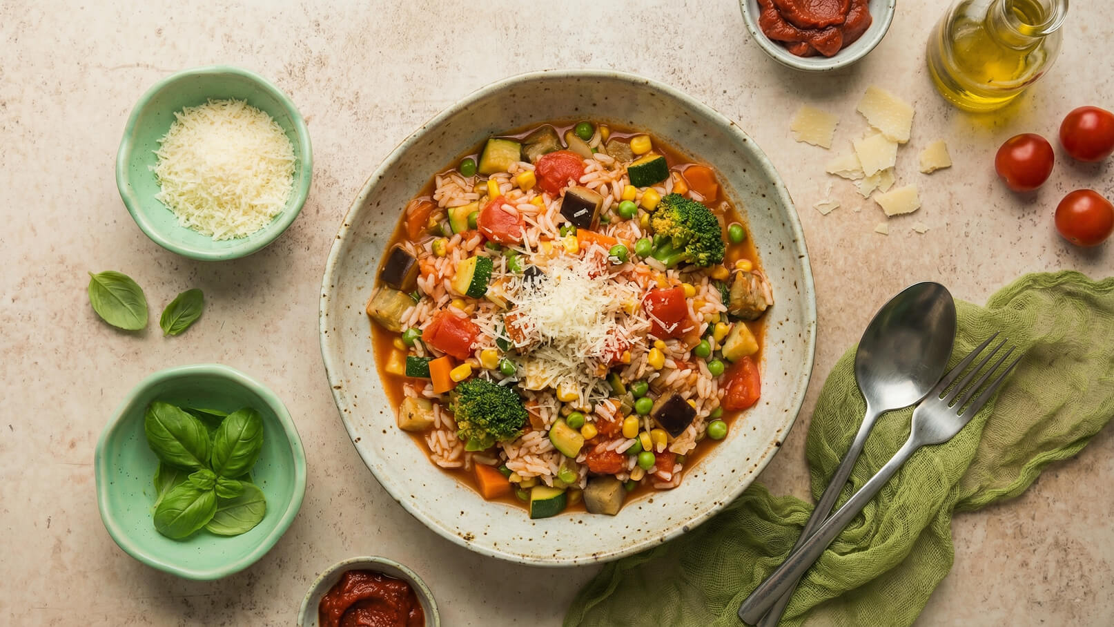 Ein tiefes Teller voller bunter Reispfanne mit Gemüse wie Brokkoli, Zucchini und Paprika, garniert mit geriebenem Käse. Neben dem Teller befinden sich Schalen mit frischem Basilikum, geriebenem Käse und Tomatensauce sowie einige Kirschtomaten. Zwei Besteckstücke liegen bereit.