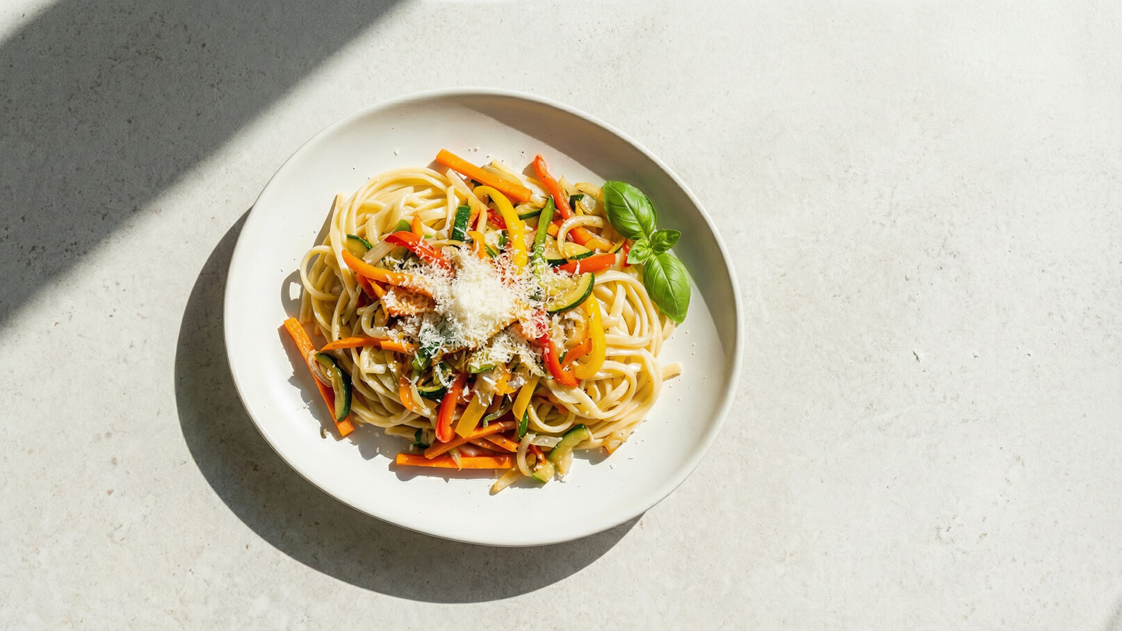 Ein Teller mit langen Nudeln, garniert mit buntem Gemüse wie Karotten, Paprika und Zucchini. Darüber liegt etwas geriebener Parmesan und ein frisches Basilikumblatt. Der Teller steht auf einer hellen, neutralen Unterlage.