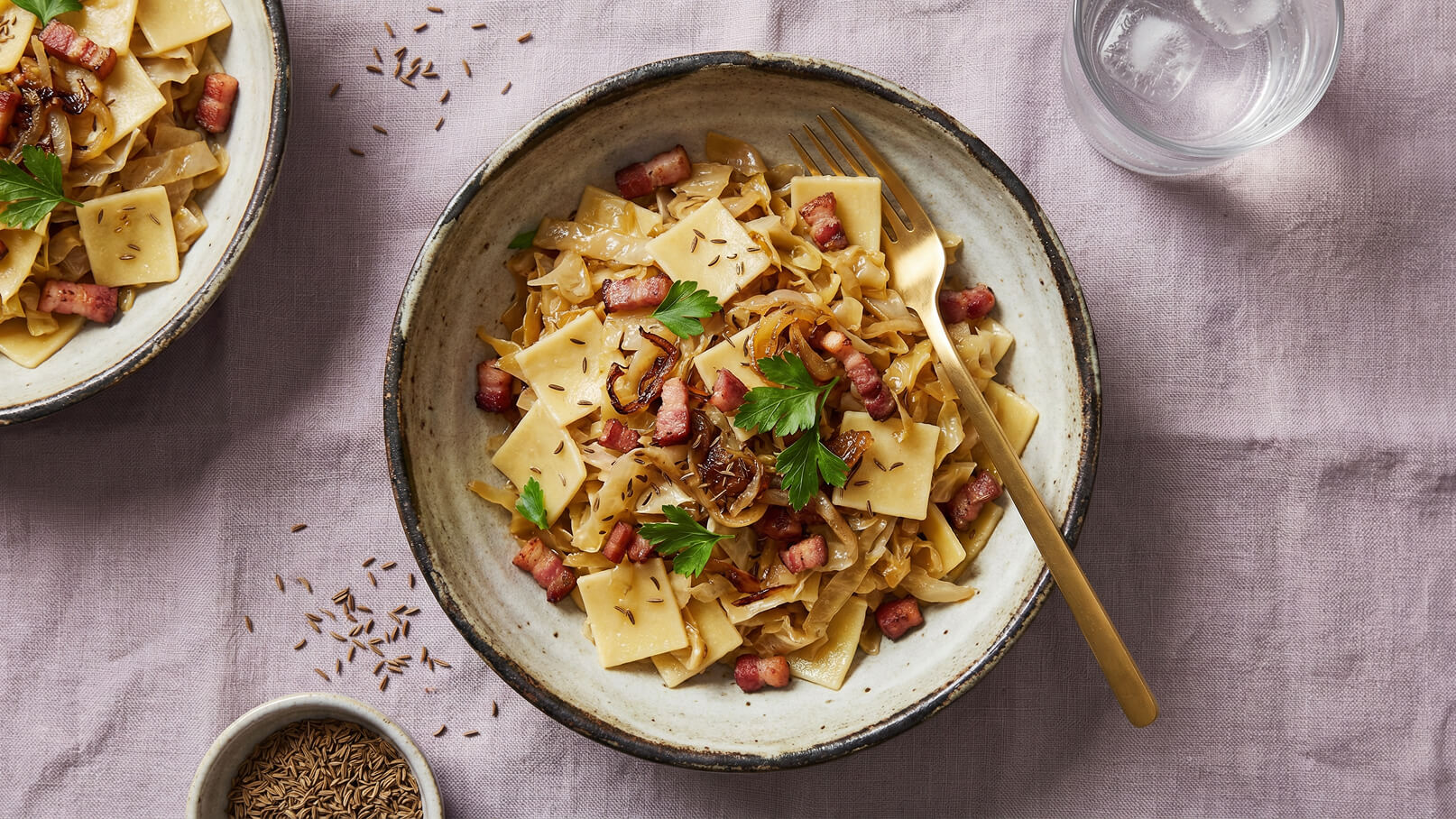 Ein tiefes Schüsselgericht mit breiten Nudeln, gebratenem Speck, Zwiebeln und Koriander. Die Nudeln sind mit Kümmelsamen bestreut, und eine goldene Gabel liegt bereit. Im Hintergrund steht ein Glas Wasser. Das Gericht ist auf einem hellen Tischcloth angerichtet.
