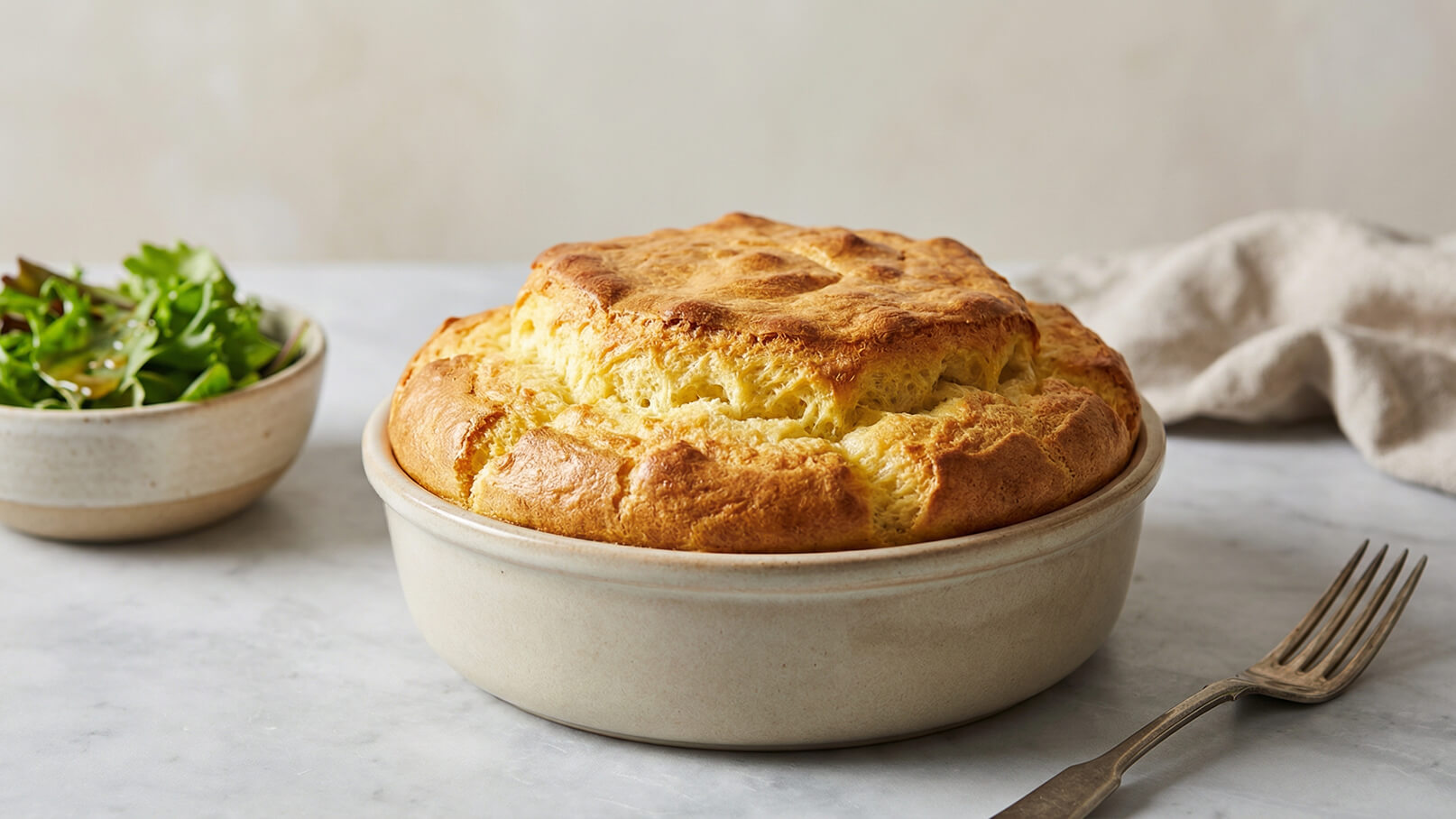 Ein goldbrauner Soufflé in einer runden Keramikschüssel steht auf einem Tisch. Nebenbei liegt eine Schüssel mit frischem Salat. Der Hintergrund ist neutral und hell, was das Gericht in den Vordergrund rückt.