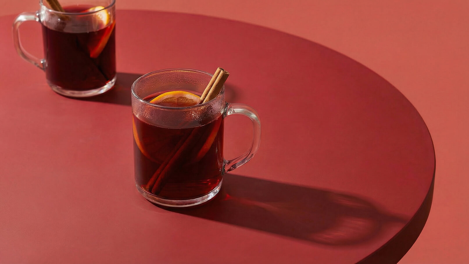 Zwei Gläser mit heißen Getränken stehen auf einem runden Tisch. Die Gläser sind transparent und enthalten eine dunkle Flüssigkeit, garniert mit einer Zimtstange und einer Orangenscheibe. Der Hintergrund und die Tischoberfläche sind in einem warmen Rotton gehalten.