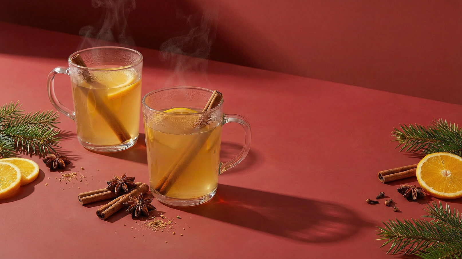 Zwei dampfende Gläser heißen Tee stehen vor einem rot Hintergrund. Jedes Glas ist mit einem Zimtstängel und einer Zitronenscheibe dekoriert. Um die Gläser sind frische Orangenscheiben, Gewürze und Tannenzweige arrangiert, die eine gemütliche Atmosphäre schaffen.