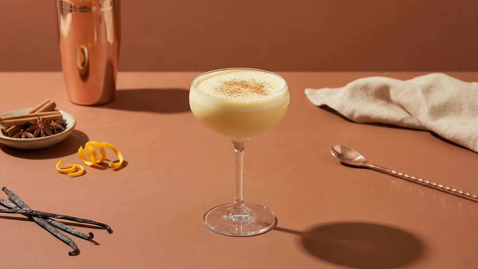Ein elegantes Glas mit einem cremigen, hellen Cocktail, garniert mit Zimt, steht auf einem braunen Tisch. Daneben liegen Zimtstangen, Sternanis, Vanilleschoten und eine Zeste aus orangener Schale. Im Hintergrund ist ein glänzender Shaker zu sehen.