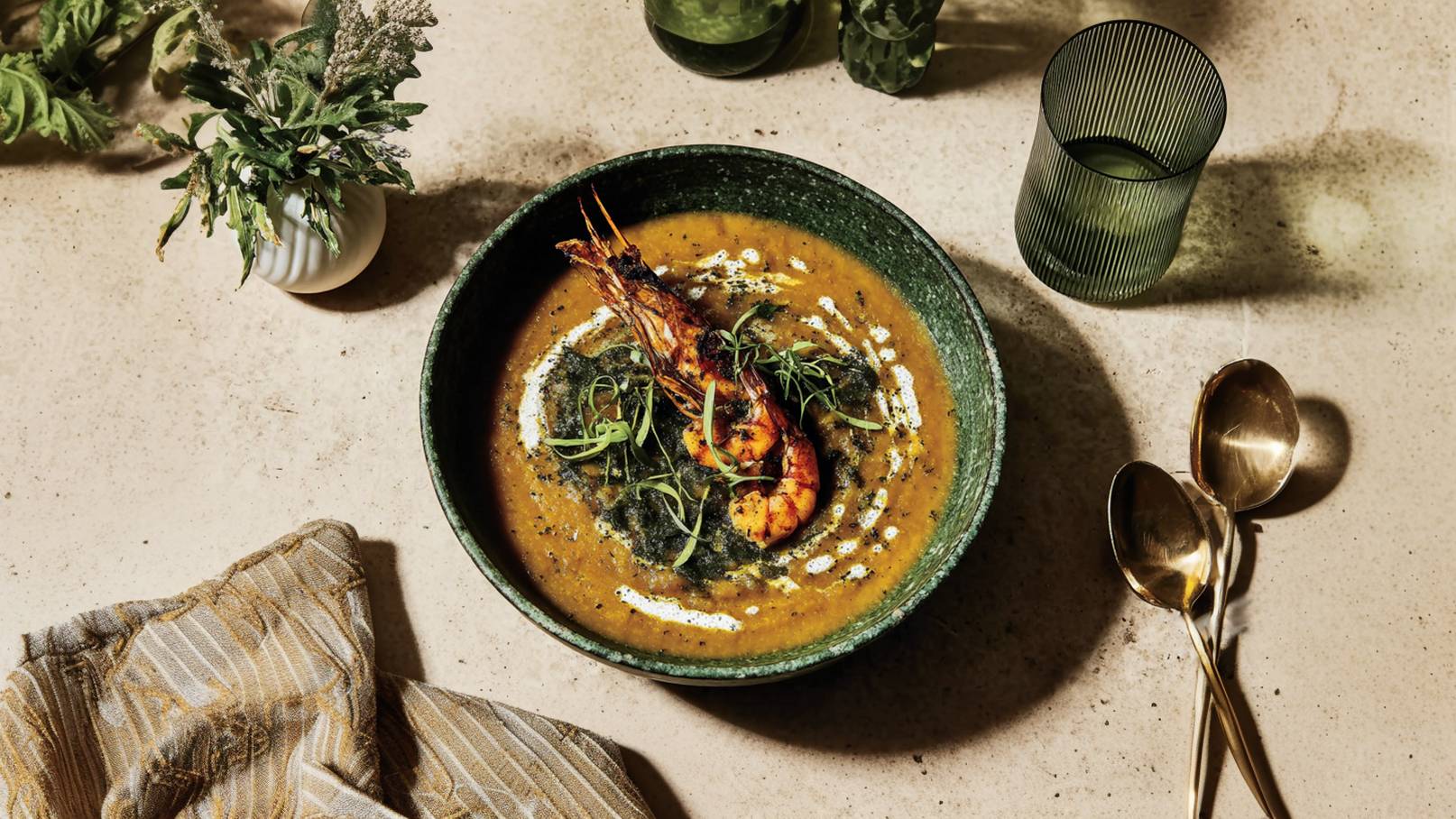 Eine grüne Schüssel enthält eine aromatische Suppe mit einer großen, gegrillten Garnele darauf. Verziert mit einer Sahnezeichnung und frischen Kräutern. Im Hintergrund sind ein längliches Tuch und goldene Löffel zu sehen.
