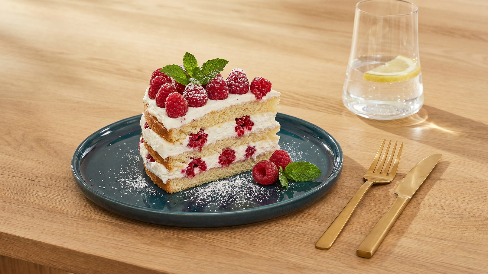 Auf einem blauen Teller liegt ein Stück Torte, geschichtet mit Sahne und Himbeeren, gekrönt von frischen Himbeeren und einem Minzblatt. Daneben sind ein Wasserglas mit Zitrone und ein Besteckset aus Gold zu sehen. Der Hintergrund ist aus Holz.