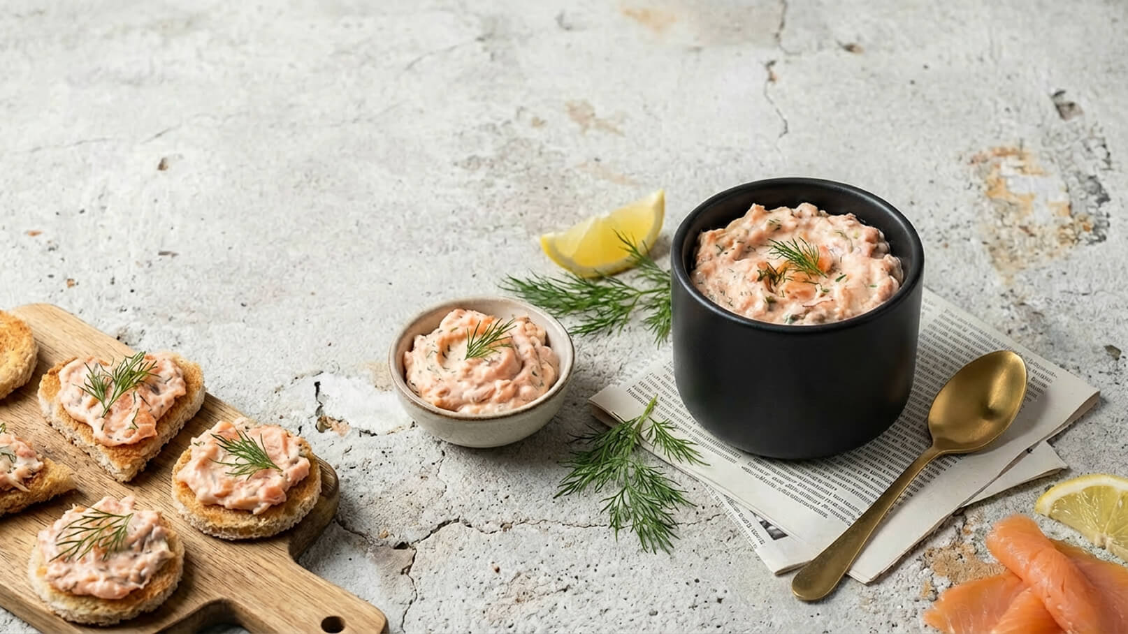 Ein schwarzer Schalenbehälter mit einer cremigen Lachs-Terrine, garniert mit Dill. Neben dem Behälter befindet sich eine kleine Schüssel mit der gleichen Mischung. Auf einem Holzbrett liegen einige kleine Brotscheiben, ebenfalls mit der Lachscreme belegt. Eine Zitronenscheibe und frischer Dill sind ebenfalls sichtbar.