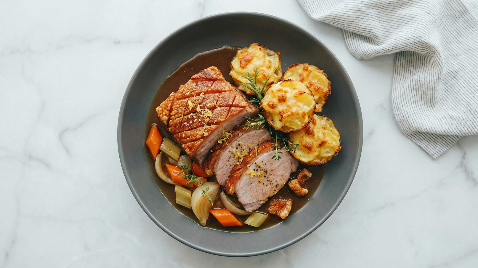 Eine Schüssel mit gebratenem Fleisch, das knusprige Haut und zarte Scheiben enthält, begleitet von Gemüse wie Karotten und Sellerie sowie Kartoffelgratin. Die Speisen sind in einer dunklen Soße angerichtet und mit frischen Kräutern garniert.