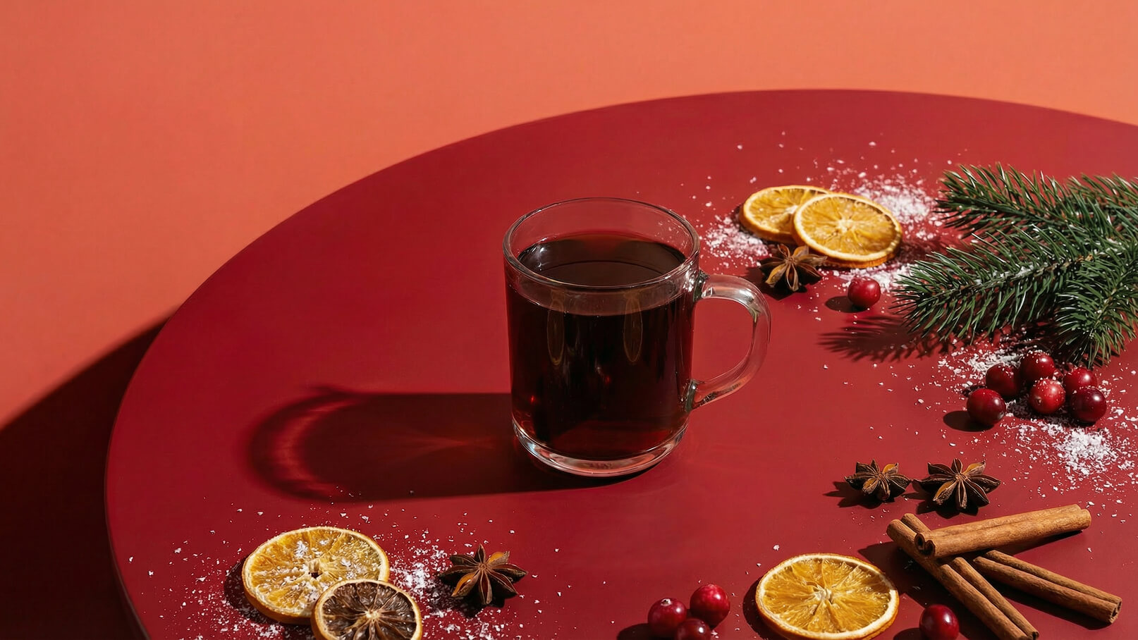 Eine gläserne Tasse mit dunklem Getränk steht auf einem roten Tisch. Um die Tasse herum sind getrocknete Orangenscheiben, Zimtstangen, Sternanis und frische Tannenzweige verteilt. Der Hintergrund ist in einem warmen Orangeton gehalten.